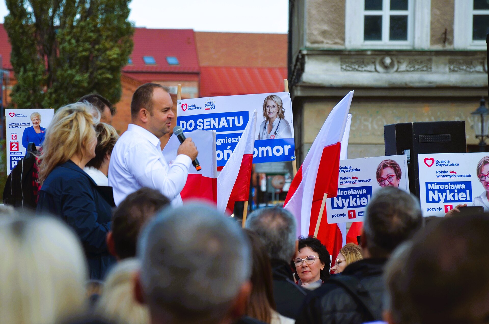 A man speaks at a Polish political rally, addressing a crowd with visible campaign signage.